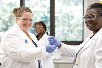 Dos científicos con batas de laboratorio y guantes sonríen y se estrechan la mano en un entorno de laboratorio.