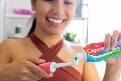 Una mujer sonríe mientras pone pasta de dientes Colgate en un cepillo de dientes en un cuarto de baño.